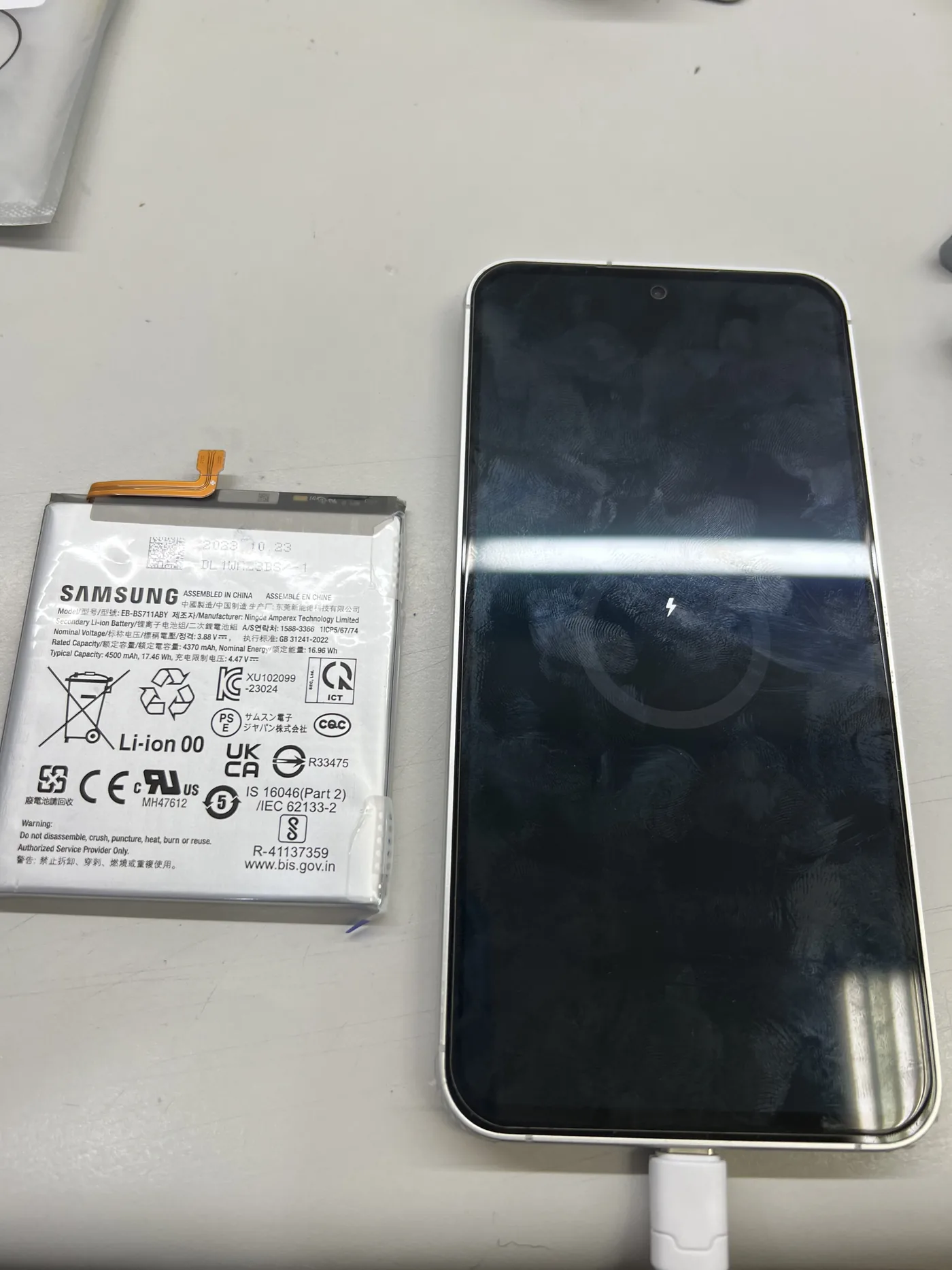 Samsung Galaxy S23 FE 換下的舊電池放在一旁，手機接上充電線後畫面顯示充電中圖示